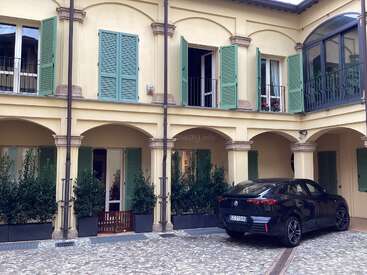 Une cour avec un bâtiment jaune, des volets verts, des plantes en pot et des passages voûtés. Une voiture noire et élégante est garée sur la surface pavée.