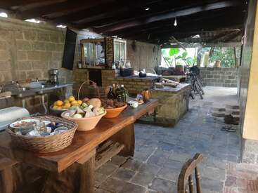 La imagen muestra una cocina rústica al aire libre con una pared de piedra, una mesa de madera y diversos utensilios de cocina, rodeada de una zona de patio con una bicicleta y otros objetos.