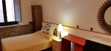 The image depicts a cozy room with a bed, table, and chairs, featuring a large window and a door, likely a living room or bedroom in a house. The walls are white, and the floor is made of wood.