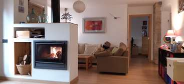 The image depicts a cozy living room with a fireplace, featuring a wooden table and chairs, a bookshelf, and a large window, all situated in a warm and inviting space.