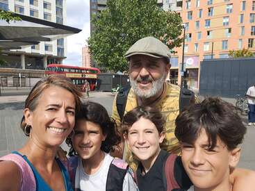 A family of five smiles for a selfie outdoors in a city. A red double-decker bus and tall buildings are visible in the background, suggesting London.