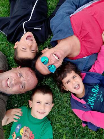 The image shows a family of five lying on the grass, smiling at the camera, with the father, mother, and three children forming a circle.