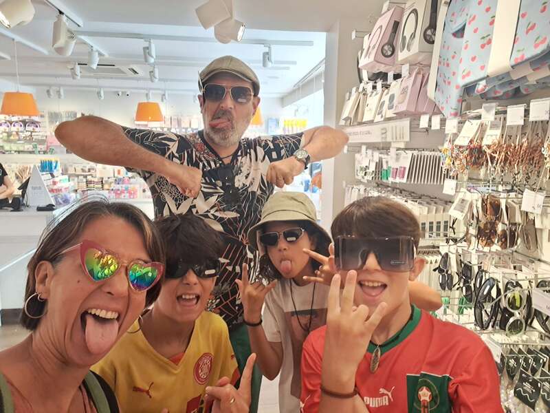 Five people posing playfully in a brightly lit store, all wearing sunglasses, making funny faces, and sticking out their tongues, exuding joyful and energetic vibes.