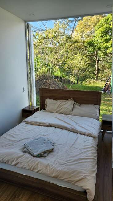 Uma cama aconchegante com lençóis bege fica ao lado de uma janela aberta, com vista para um jardim verdejante. Toalhas dobradas repousam sobre a cama, convidando ao relaxamento.