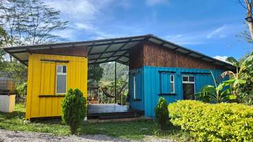 This image shows a small house made from yellow and blue shipping containers, surrounded by greenery, with a hammock in the open central area, under clear skies.