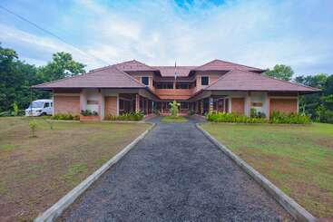 The image depicts a two-story building with a red roof, featuring a gravel path leading to the entrance, surrounded by lush greenery and a serene blue sky.