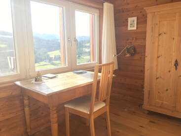 The image depicts a cozy room with a desk, chair, and armoire, featuring a large window with a view of the outdoors, all crafted from light-colored wood.