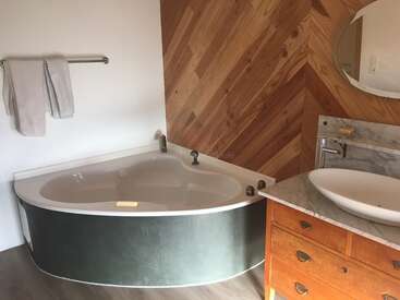 The image depicts a bathroom with a corner bathtub, a sink, and a wooden vanity, featuring a white marble countertop and a round mirror on the wall.