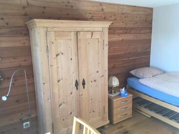 The image depicts a cozy room with a wooden wardrobe, nightstand, and bed, featuring a wooden plank wall and floor, with a white ceiling and a small chair in the foreground.