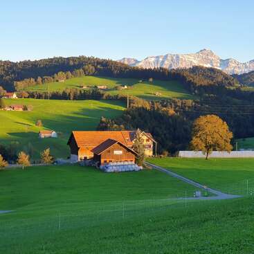 The image depicts a serene rural landscape featuring a large barn, rolling hills, and a majestic mountain range in the background under a clear blue sky.