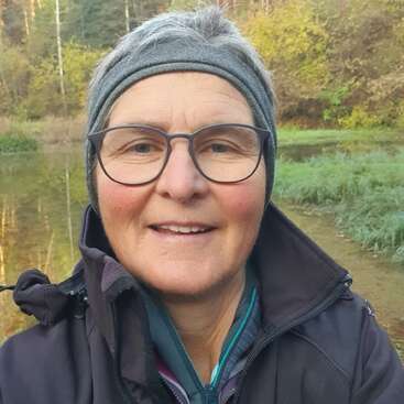 Une femme aux cheveux gris courts, portant des lunettes et un bandeau gris, sourit en plein air. Elle porte une veste noire et se tient au bord d'une rivière dans une forêt.