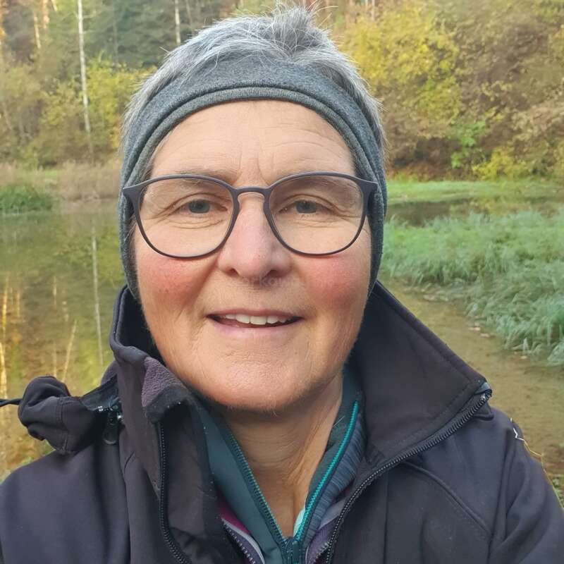 Una mujer de pelo corto y gris, con gafas y una cinta gris en la cabeza, sonríe al aire libre. Lleva una chaqueta negra, de pie junto a un río en un entorno forestal.