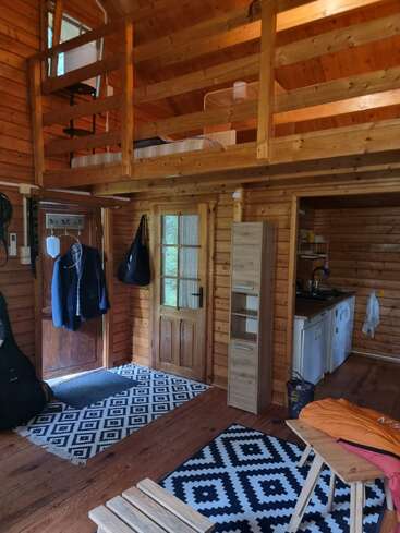 Esta acogedora cabaña de madera cuenta con una cama en altillo, una pequeña zona de cocina, alfombras estampadas, muebles sencillos, abrigos colgantes y un ambiente acogedor y rústico con luz natural.