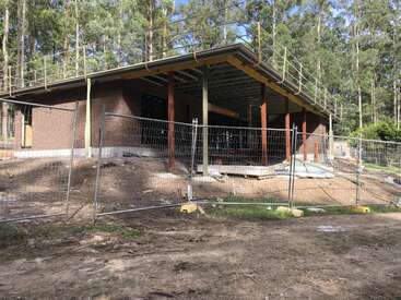 This image shows a partially constructed brick building surrounded by a temporary metal fence, located in a forested area with scattered construction materials and dirt.