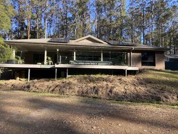 Esta imagem mostra uma casa moderna com uma grande varanda envidraçada, painéis solares no telhado, construída sobre palafitas e cercada por árvores em uma área arborizada.