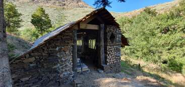Esta imagen muestra una pequeña cabaña rústica construida con piedras y troncos apilados, situada en una zona pintoresca, montañosa y boscosa, bajo un cielo azul brillante.