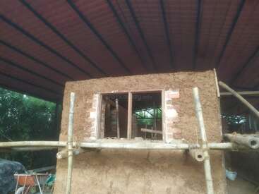 This image shows an unfinished earthen house with a brick-framed window. Bamboo scaffolding surrounds the building, and a metal roof provides shelter over the construction site.