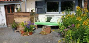 Un acogedor patio ajardinado presenta un banco blanco con cojines verdes y almohadas, cajas de madera, flores de colores en macetas, flores amarillas y una ventana con contraventanas abiertas.
