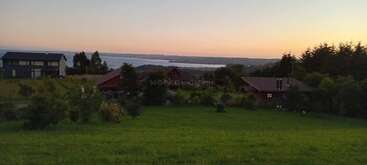 Este sereno paisaje muestra tres casas rodeadas de exuberante vegetación, árboles y un espacioso césped, con una vista lejana de un tranquilo lago al atardecer.