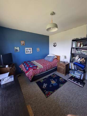 La habitación tiene una cama individual con ropa de cama a rayas, una pared azul, una estantería, una mesita de noche, una alfombra con motivos de robots, un escritorio, una lámpara y cuadros en la pared.