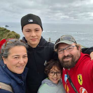 A happy family of four poses for a selfie outdoors, with the ocean and cliffs in the background under a cloudy sky, all smiling closely together.