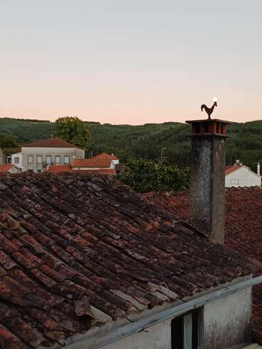 Dieses Foto zeigt rustikale rote Ziegeldächer bei Sonnenuntergang, einen Schornstein mit der Silhouette eines Hahns und üppig grüne Hügel im Hintergrund, die an ländliche Ruhe erinnern.