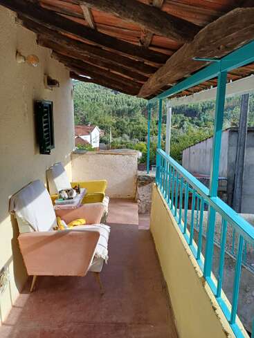 Dieser gemütliche Balkon ist mit zwei alten Stühlen, einem kleinen Tisch mit Obst, rustikalen Holzbalken und einem blauen Geländer ausgestattet und bietet einen Blick auf das üppige Grün und die nahegelegenen Dorfhäuser.