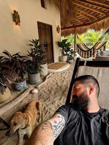 Un homme barbu se détend sur une véranda sablonneuse, aux côtés d'un chien brun. Des plantes en pot, des hamacs, un toit de chaume et des portes en bois créent une atmosphère paisible et tropicale.