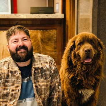 Un homme barbu vêtu d'une veste à carreaux sourit, assis à côté d'un gros chien brun au pelage soyeux. Ils semblent à l'aise et heureux à l'intérieur, près d'un comptoir.