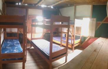 Cette image montre une pièce en bois avec trois ensembles de lits superposés, plusieurs matelas, des sols et des murs en bois, éclairée par la lumière naturelle du soleil qui passe par les fenêtres.