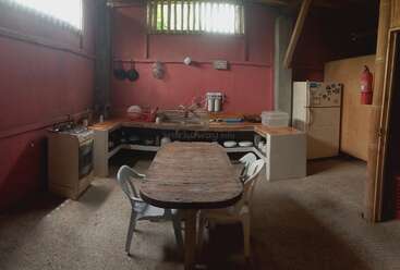 Cette cuisine rustique présente des murs rouges, une table en bois avec des chaises en plastique blanc, des étagères ouvertes, une cuisinière à gaz, un réfrigérateur, des casseroles suspendues et une petite fenêtre pour l'aération.