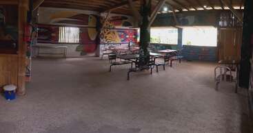 Cette salle à manger en plein air est ornée de peintures murales colorées représentant des oiseaux et des animaux, de poutres en bois, de tables et de chaises simples et de grandes fenêtres laissant entrer la lumière naturelle.