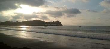 Une belle plage au coucher du soleil avec des vagues douces qui roulent doucement sur le rivage, des bateaux au loin, des collines silhouettées et un ciel rempli de nuages dramatiques et dispersés.