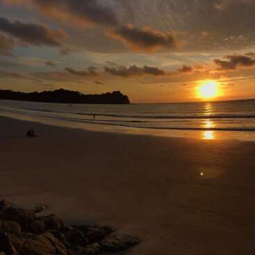 L'image représente une scène de plage sereine au coucher du soleil, avec un ciel vibrant parsemé de nuages, un océan calme et un rivage sablonneux avec des rochers au premier plan.