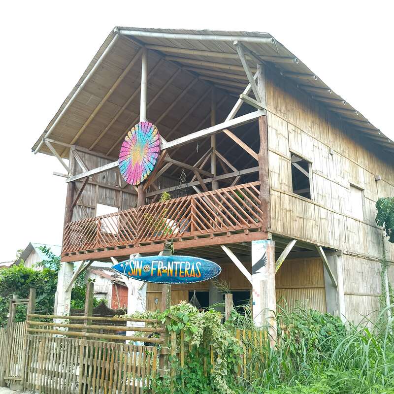 Cette maison rustique en bambou de deux étages est dotée d'un balcon ouvert, d'œuvres d'art colorées et d'une planche de surf portant la mention "Sin Fronteras". Elle est entourée de verdure et d'une clôture en bois.