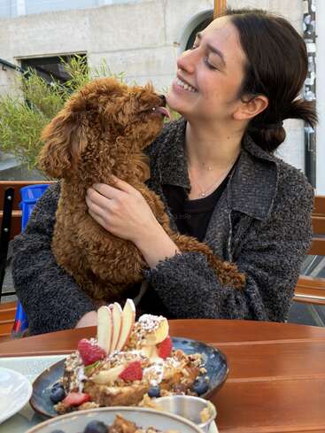 Eine Frau lächelt liebevoll, während sie einen flauschigen braunen Hund im Arm hält, der ihr Gesicht ableckt. Auf dem Tisch stehen Teller mit Waffeln, Obst und Sahne.