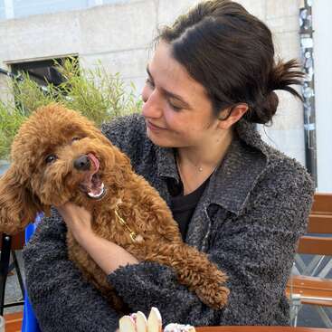 Eine Frau in einer grauen Jacke hält liebevoll einen flauschigen braunen Hund im Arm, der eine lustige Grimasse schneidet – draußen vor einem Café mit Holztischen.