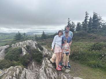 A family of four and their dog stand on a rocky hilltop surrounded by greenery and trees, with a scenic, cloudy rural landscape in the background.