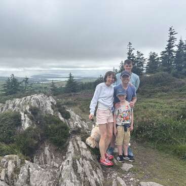 Uma família de quatro pessoas, com um cachorrinho, posa no topo de uma colina rochosa. O campo verde, as árvores e o céu nublado criam um cenário pitoresco e tranquilo ao ar livre.