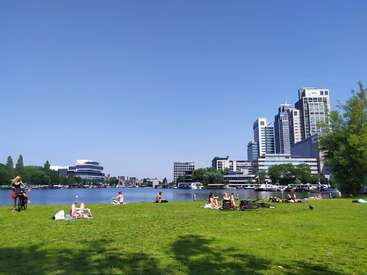 The image depicts a serene park scene with a group of people relaxing on the grass, set against a backdrop of a large body of water and a city skyline.