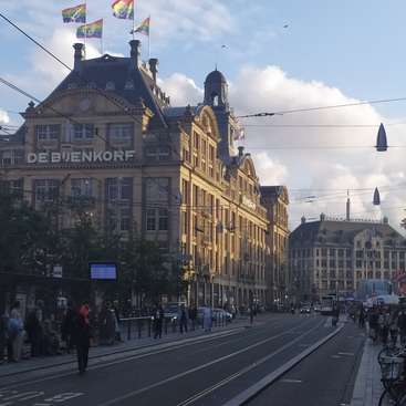 The image depicts a city street with a large building in the background, featuring flags on top and a tram track running down the center of the road.