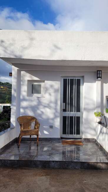 A small, white-walled porch features a wicker chair, tiled floor, sliding door, potted plant, lantern light, and a doormat under a partly cloudy sky.
