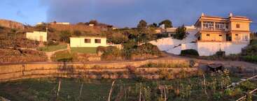 A terraced hillside with lush gardens, small, simple buildings, and a large house. Sunlight bathes the area, casting warm hues over the serene landscape.