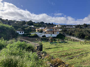 A rural landscape with a yellow house, green fields, garden plots, and a playground. Hills, trees, and scattered houses are beneath a mostly sunny blue sky.