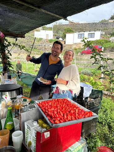 A smiling man and woman stand outdoors, preparing food by a table covered with fresh strawberries, drinks, and supplies. A relaxed, cheerful atmosphere with greenery around.