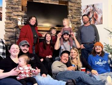 Une joyeuse famille se réunit dans un salon confortable, souriante et détendue. Enfants et adultes posent ensemble devant une cheminée rustique en pierre et un téléviseur.