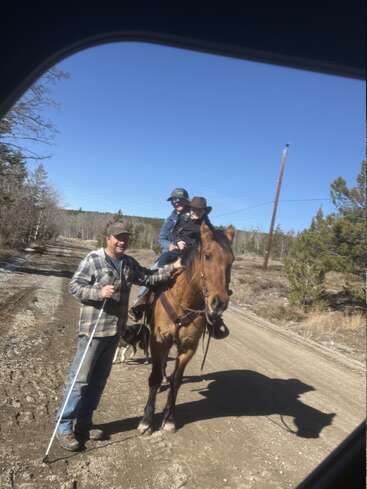 Three people, two wearing cowboy hats, are outdoors on a dirt road. One person leads a horse carrying two riders. It’s a sunny, clear day with trees surrounding.