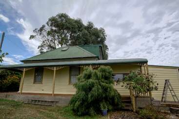 A yellow house with a green roof and verandah sits beneath a cloudy sky, surrounded by trees, shrubs, a ladder, and a potted plant in the yard.