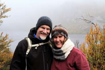 A smiling couple dressed warmly in hats and scarves stands outdoors on a misty, foggy day near a body of water, enjoying the chilly weather together.