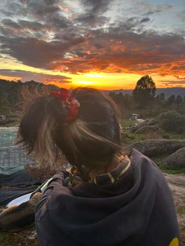 Une personne portant une queue de cheval et un chouchou rouge est assise en plein air, face à un ciel crépusculaire aux couleurs vives, parsemé de nuages, d'arbres et de rochers, avec un chien qui se repose à proximité.